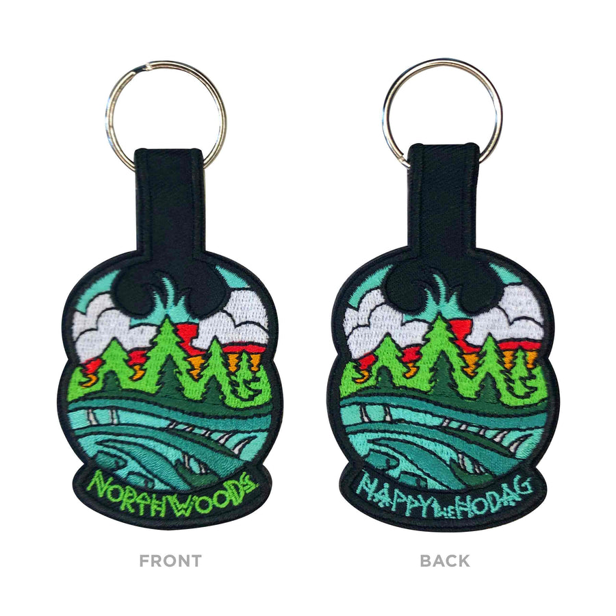 black keychains with colorful embroidery of sky, trees, water and a Meshipeshu hiding in the waves. Front has text Northwoods, back has text Happy the Hodag