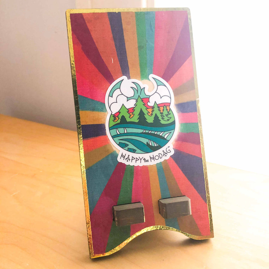 Sticker shaped like Happy the hod's head with colorful artwork of sky, trees, water and a Meshipeshu hiding in the waves with the text Happy the Hodag shown applied to a colorful smart phone holder